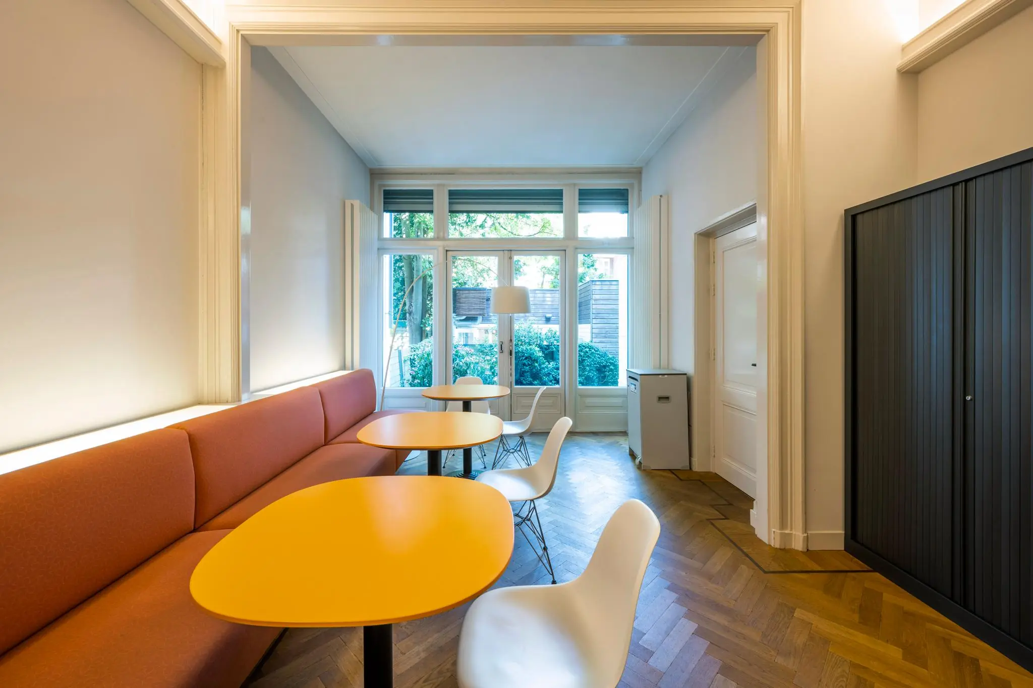 Moderne zithoek in een lichte ruimte aan de Paulus Potterstraat met oranje tafels, witte stoelen en een lange rode bank.